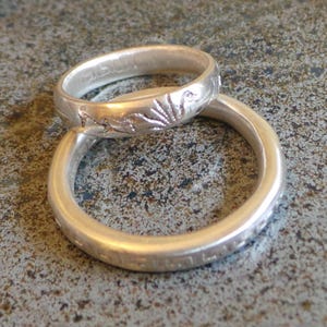May include: Two silver rings, one stacked on top of the other. The top ring has a floral design and the bottom ring is plain. The rings are on a speckled gray surface.