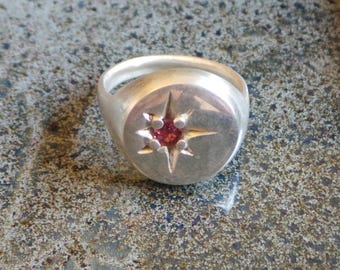 May include: A silver signet ring featuring a circular face with a polished finish. In the center of the face is a small, round, pink gemstone set within a four-pointed star engraving that radiates outward from the stone.