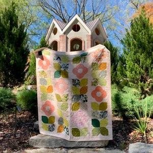 May include: A pink and green floral quilt with a white background. The quilt is hanging on a stone wall in front of a small wooden structure.