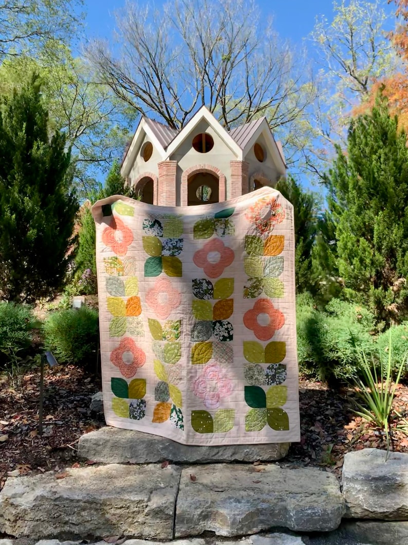 May include: A pink and green floral quilt with a white background. The quilt is hanging on a stone wall in front of a small wooden structure.
