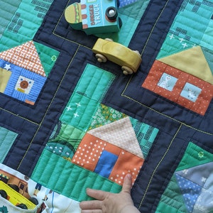 Op de afbeelding: Een groene en blauwe patchworkdeken met een verscheidenheid aan kleurrijke vierkanten van stof. Een kinderhand reikt naar de deken, waarop een speelgoedtruck en een speelgoedauto te zien zijn. De deken is perfect voor een kinderkamer of slaapkamer.