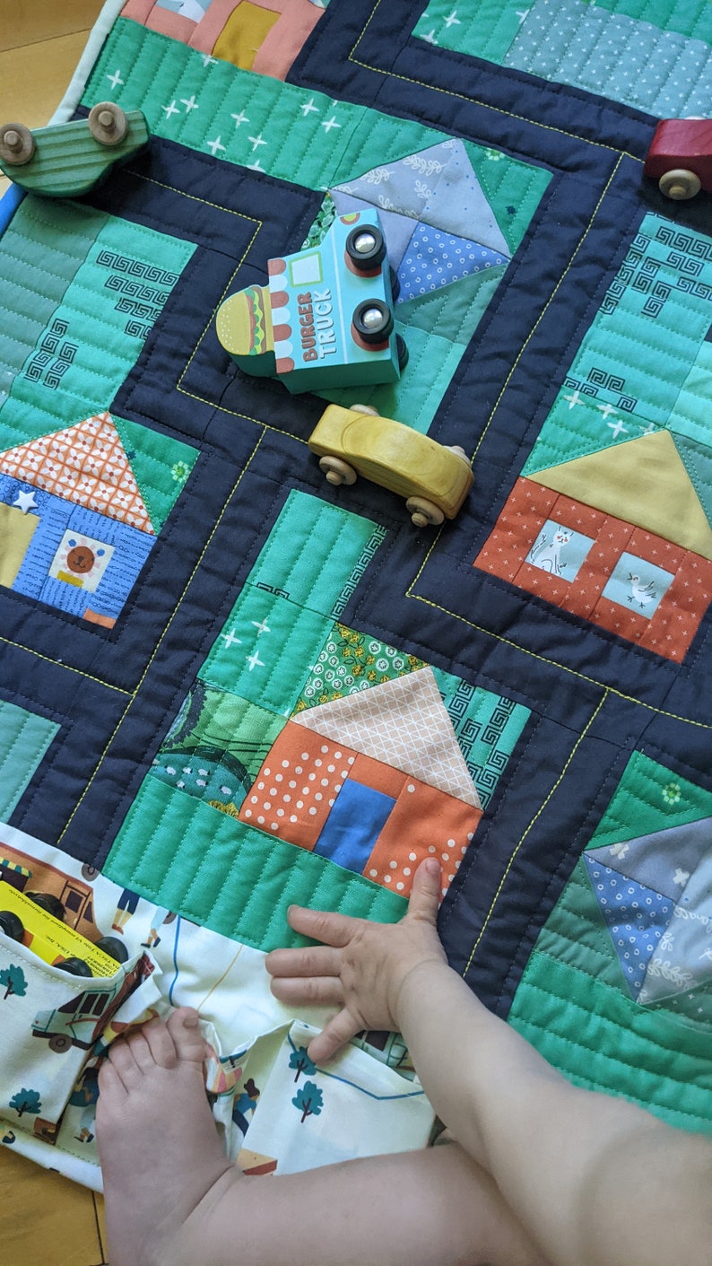 Op de afbeelding: Een groene en blauwe patchworkdeken met een verscheidenheid aan kleurrijke vierkanten van stof. Een kinderhand reikt naar de deken, waarop een speelgoedtruck en een speelgoedauto te zien zijn. De deken is perfect voor een kinderkamer of slaapkamer.