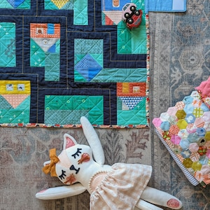 Op de afbeelding: Een kleurrijk patchwork-deken met een geometrisch ontwerp in tinten blauw, groen en geel. Een wit knuffeldier met een roze jurk en bruin haar ligt op het deken. Een kleine speelgoedauto is zichtbaar in de rechterbovenhoek van de afbeelding.