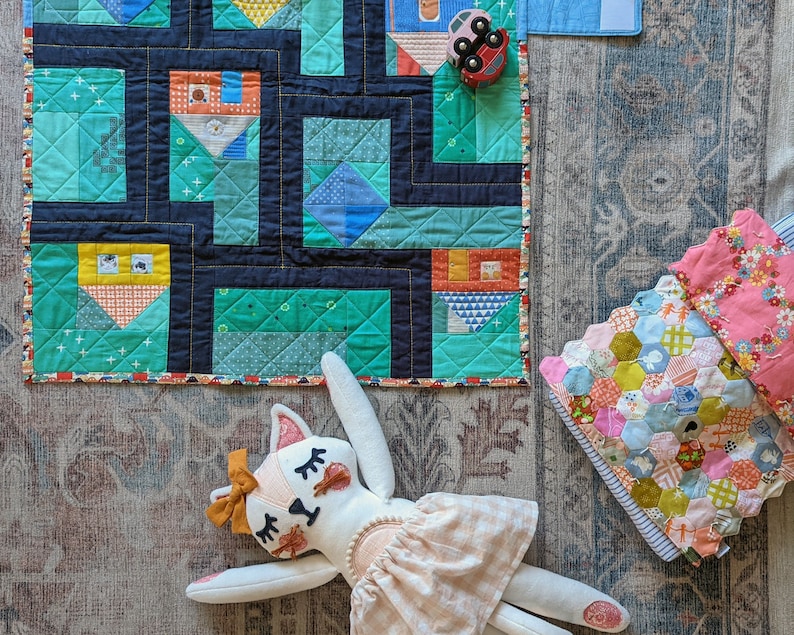 Op de afbeelding: Een kleurrijk patchwork-deken met een geometrisch ontwerp in tinten blauw, groen en geel. Een wit knuffeldier met een roze jurk en bruin haar ligt op het deken. Een kleine speelgoedauto is zichtbaar in de rechterbovenhoek van de afbeelding.