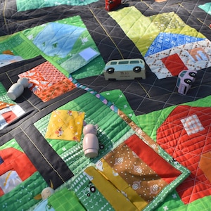 Op de afbeelding: Een kleurrijke speelmat met een patchwork-ontwerp met huizen, schuren en wegen. De mat is gemaakt van stof en heeft een gestikte textuur. Er zijn houten speelgoedauto's en poppetjes op de mat.