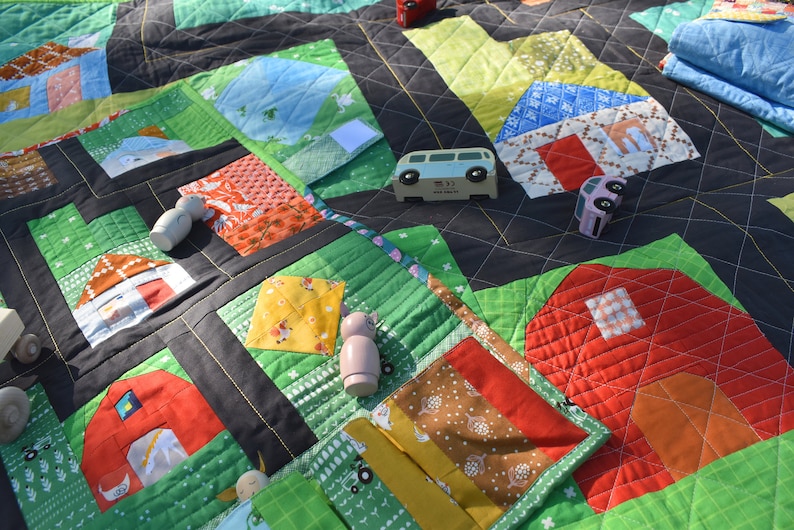 Op de afbeelding: Een kleurrijke speelmat met een patchwork-ontwerp met huizen, schuren en wegen. De mat is gemaakt van stof en heeft een gestikte textuur. Er zijn houten speelgoedauto's en poppetjes op de mat.