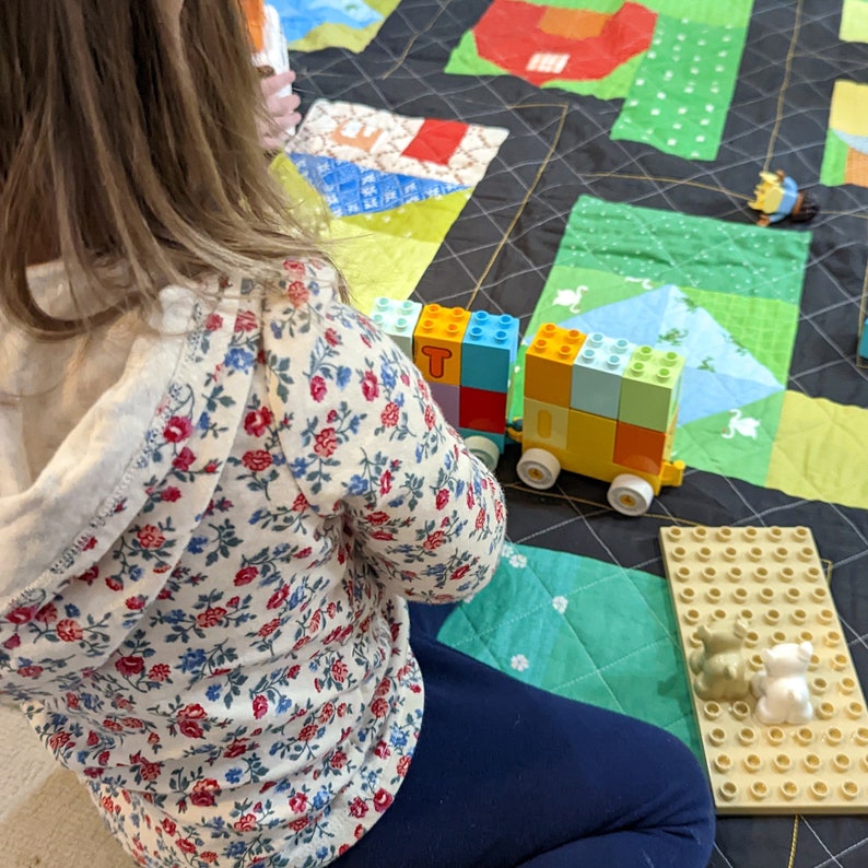 Op de afbeelding: Een kind speelt met een kleurrijke speelgoedtrein gemaakt van bouwstenen op een zwart-witte patchworkdeken. De trein is gemaakt van gele, blauwe, rode en groene blokken. De deken heeft verschillende patronen, waaronder bloemen, sterren en geometrische vormen.