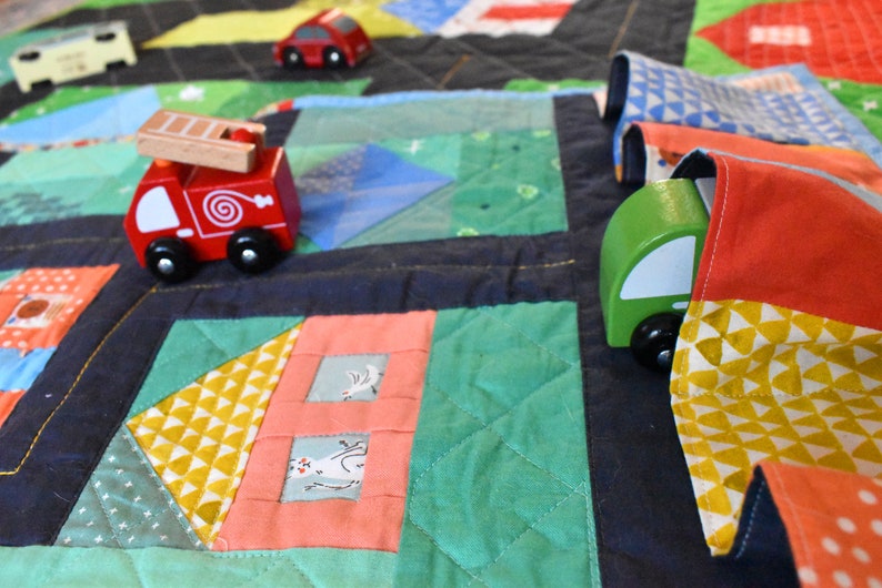 Op de afbeelding: Een kleurrijke speelmat met een groen, rood en blauw patchwork ontwerp. Een rode brandweerwagen, een groene auto en een blauwe auto staan op de mat. De mat is gemaakt van stof en heeft een gestikte textuur.