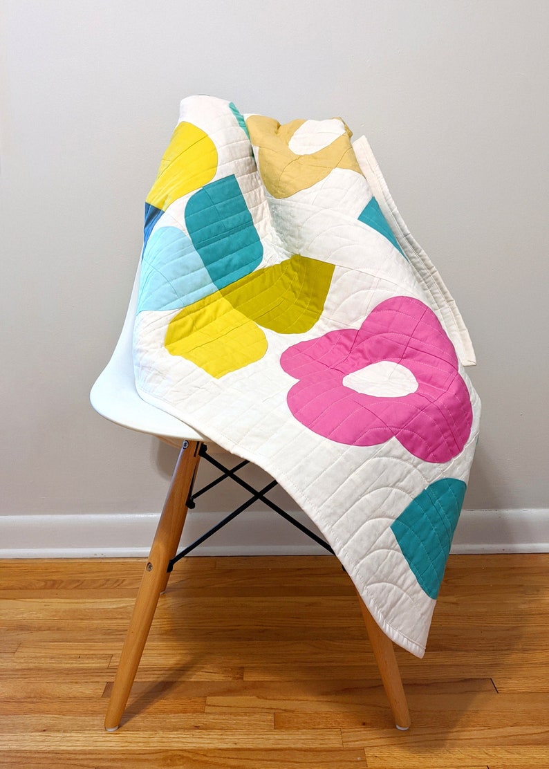 May include: A white quilt with a geometric pattern of pink, yellow, teal, and blue shapes. The quilt is folded and draped over a white chair with wooden legs.