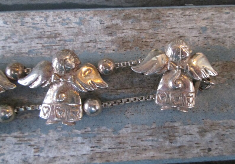 May include: Silver-toned angel bracelet featuring detailed angel figures with wings and heart-shaped accents. The bracelet includes small, round beads and a delicate chain, perfect for adding a touch of charm to any outfit.