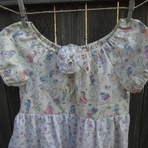 May include: A cream-colored dress with short sleeves and a floral pattern. The bodice features a print of figures and letters, with a white floral accent at the neckline. The skirt has a floral print. The dress is hanging on a clothesline.