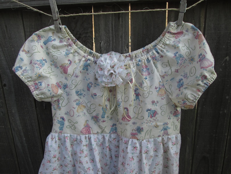 May include: A cream-colored dress with short sleeves and a floral pattern. The bodice features a print of figures and letters, with a white floral accent at the neckline. The skirt has a floral print. The dress is hanging on a clothesline.