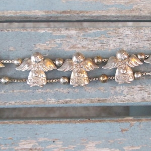 May include: A silver-toned bracelet featuring six angel charms with detailed wings and beaded accents. The bracelet has a toggle clasp and is displayed on a weathered, light blue wooden surface. The angels are evenly spaced along the chain.