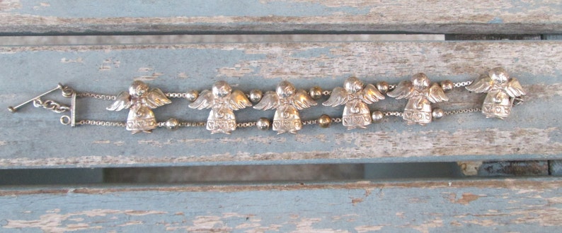 May include: A silver-toned bracelet featuring six angel charms with detailed wings and beaded accents. The bracelet has a toggle clasp and is displayed on a weathered, light blue wooden surface. The angels are evenly spaced along the chain.