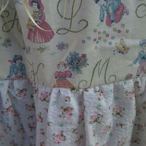 May include: A close-up of a fabric with a vintage design. The top portion features illustrations of children and floral patterns. The bottom has a white fabric with pink floral patterns. The fabric is likely used for a decorative item.
