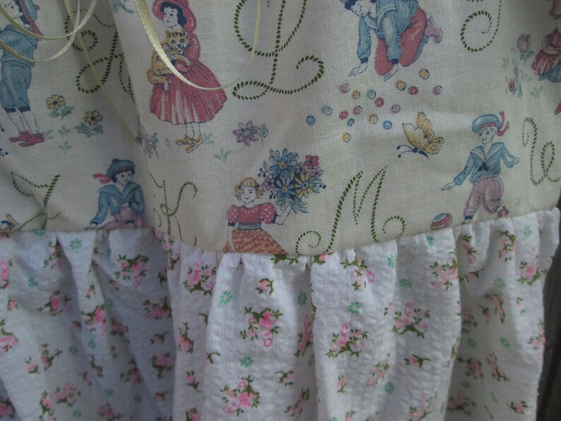 May include: A close-up of a fabric with a vintage design. The top portion features illustrations of children and floral patterns. The bottom has a white fabric with pink floral patterns. The fabric is likely used for a decorative item.