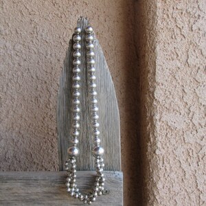 May include: A silver beaded necklace with varying bead sizes hangs against a weathered wooden surface. The necklace has a double strand design at the bottom and a simple clasp.