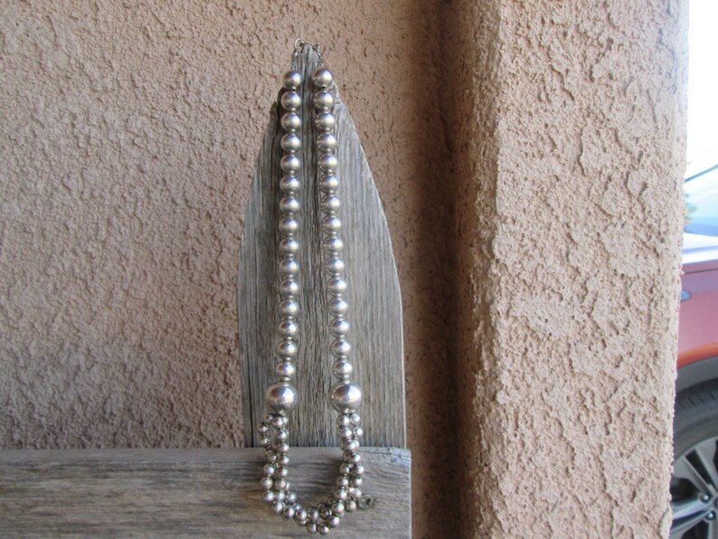 May include: A silver beaded necklace with varying bead sizes hangs against a weathered wooden surface. The necklace has a double strand design at the bottom and a simple clasp.