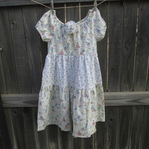 May include: A cream-colored dress with a floral and figure pattern, hanging on a clothesline. The dress features short sleeves, a tiered skirt, and a decorative flower at the neckline. The fabric has a light, airy appearance, suggesting a summer garment.