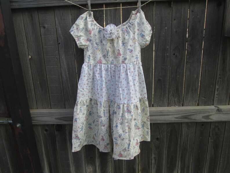 May include: A cream-colored dress with a floral and figure pattern, hanging on a clothesline. The dress features short sleeves, a tiered skirt, and a decorative flower at the neckline. The fabric has a light, airy appearance, suggesting a summer garment.