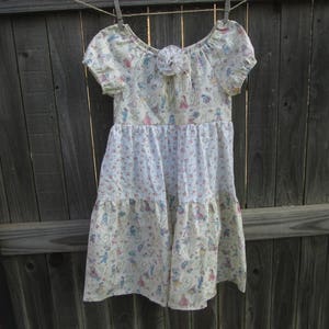 May include: A cream-colored dress with short sleeves and a tiered skirt, hanging on a clothesline. The dress features a floral and figure pattern in pink, blue, and green. A decorative flower is at the neckline.