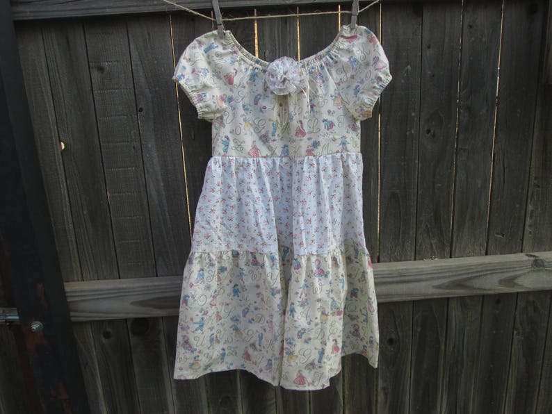 May include: A cream-colored dress with short sleeves and a tiered skirt, hanging on a clothesline. The dress features a floral and figure pattern in pink, blue, and green. A decorative flower is at the neckline.