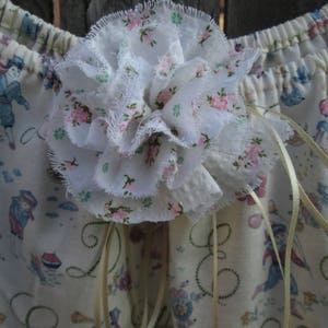 May include: A white fabric flower with frayed edges is the focal point, adorned with pink floral accents. The background features a cream-colored fabric with a repeating pattern of figures in blue and pink, and decorative cursive script.