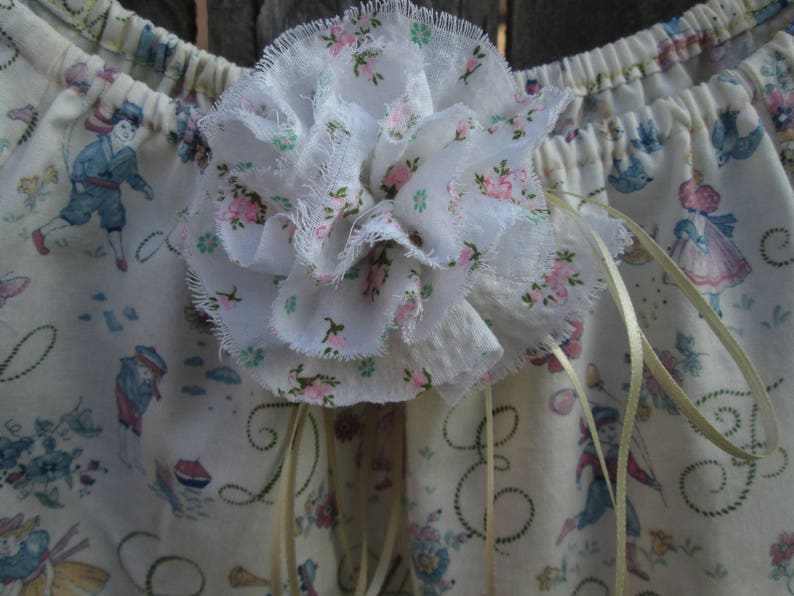 May include: A white fabric flower with frayed edges is the focal point, adorned with pink floral accents. The background features a cream-colored fabric with a repeating pattern of figures in blue and pink, and decorative cursive script.