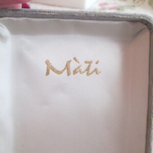 May include: An open jewellery box with a grey velvet exterior and a white satin interior. The box has the word "Mati" printed in gold lettering on the inside.