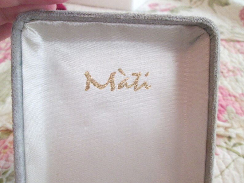 May include: An open jewellery box with a grey velvet exterior and a white satin interior. The box has the word "Mati" printed in gold lettering on the inside.