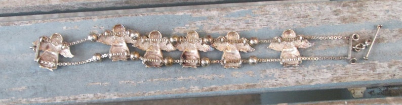 May include: A silver-toned bracelet featuring six angel charms with outstretched wings. The bracelet has a delicate chain design with small beads between the angel figures. The clasp is a simple bar and loop closure.