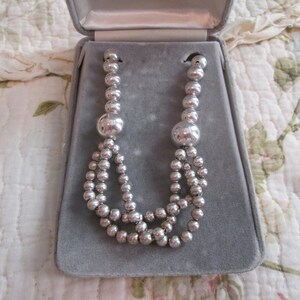 May include: A silver necklace featuring multiple strands of small, round beads and two larger, spherical beads. The necklace is presented in an open, grey velvet jewellery box. The box's interior provides a soft backdrop for the necklace's design.