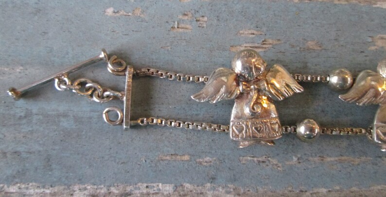 May include: Silver-toned angel bracelet with a toggle clasp. The bracelet features small angel charms with detailed wings and heart-shaped accents on their gowns. The chain is a delicate box chain style.