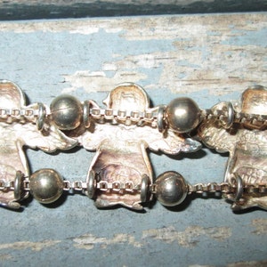 May include: Silver-toned angel bracelet with detailed angel figures and round beads. The angels are connected by a delicate chain. The bracelet is displayed on a weathered, light blue surface.