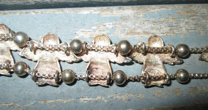 May include: Silver-toned angel bracelet with detailed angel figures and round beads. The angels are connected by a delicate chain. The bracelet is displayed on a weathered, light blue surface.