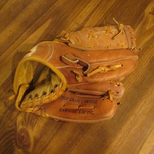 May include: A brown leather baseball glove with white stitching. The glove has the text "Top Grain Cowhide" and "Rawhide Lacing" printed on it.