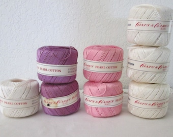 May include: A collection of crochet thread spools in various colors, including white, pink, and purple. The spools are labeled "Pearl Cotton" and "Coats & Clark's". The image showcases the thread's texture and color variations, ideal for crafting projects.