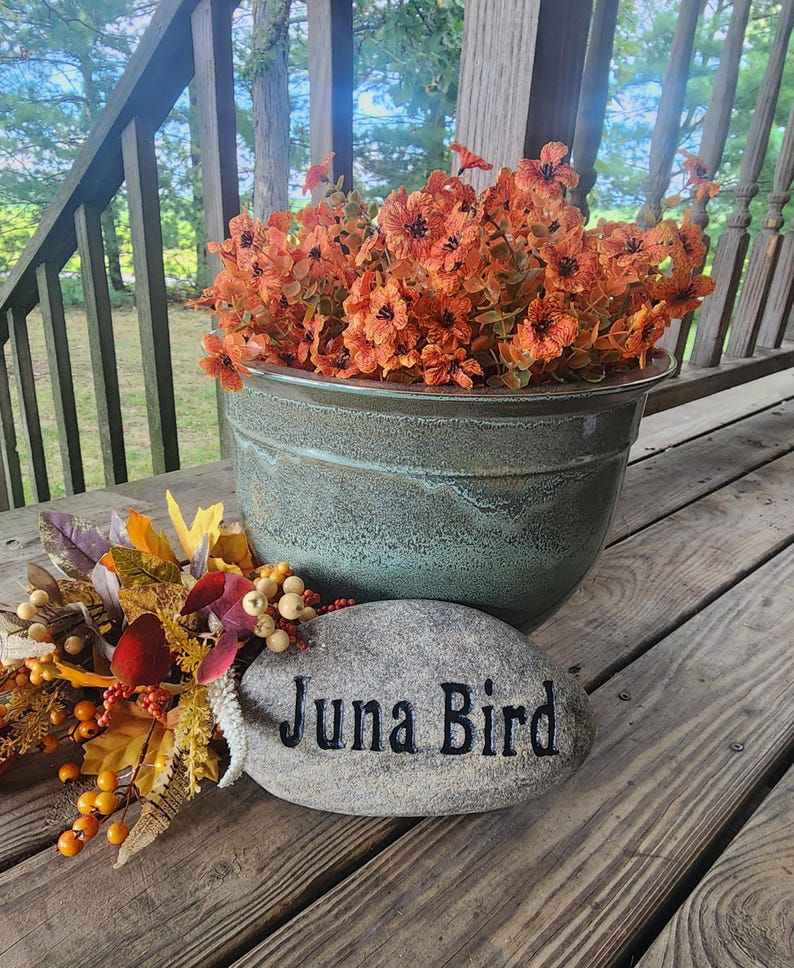 May include: A green ceramic planter with orange flowers and a gray stone with the text "Juna Bird" on it.