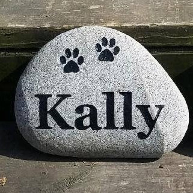 May include: A gray stone with two black paw prints and the name "Kally" written in black lettering.