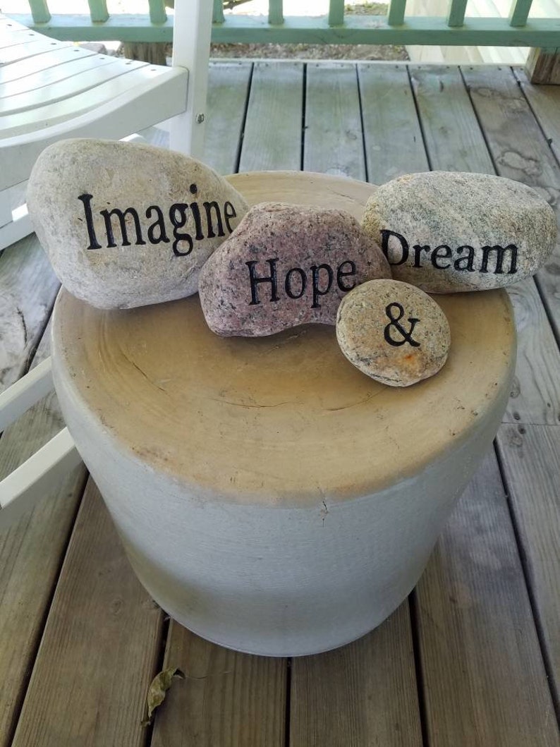 May include: Four painted stones with the words "Imagine", "Hope", "Dream", and "&" on a light brown wooden surface.