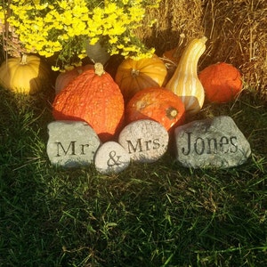 May include: Two gray stones with the words "Mr. & Mrs. Jones" engraved on them. The stones are surrounded by gourds and fall foliage.