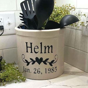 May include: A white ceramic crock with black lettering that reads "Helm Jan. 26, 1985" and a black heart with vines. The crock is filled with black kitchen utensils and green plants.