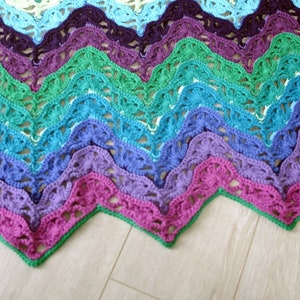 May include: A colorful crochet blanket with a chevron pattern in shades of green, blue, purple, and pink. The blanket is made with a delicate lace stitch and has a scalloped edge.