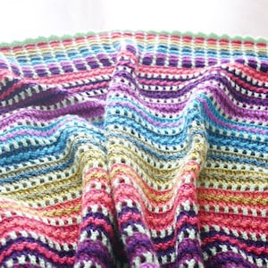 May include: A colorful striped crochet blanket with a textured pattern. The blanket is made with a variety of colors, including blue, purple, pink, and yellow. The blanket is folded in half and the edges are visible. The text "Felted Button" is visible in the bottom right corner of the image.