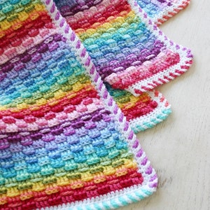 May include: A rainbow crochet baby blanket with a white and purple border. The blanket is folded in thirds, showing the different colors of the rainbow.