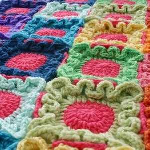 Può includere: Una coperta all'uncinetto colorata fatta di quadrati con bordi arricciati. I quadrati sono in tonalità di rosa, blu, verde, giallo e arancione.
