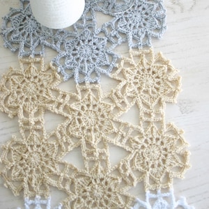 May include: A crocheted table runner with a repeating pattern of snowflakes. The runner is made in three colors: white, light beige, and light blue.