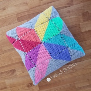 May include: A colorful crocheted pillow with a geometric pattern in shades of gray, pink, purple, blue, green, and yellow. The pillow is on a wooden floor.