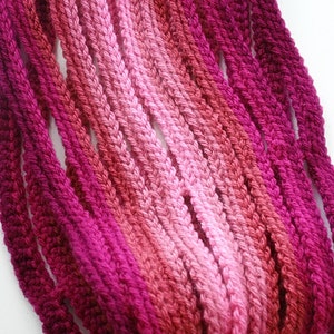 May include: A collection of pink, red, and purple yarn strands in various shades, arranged in a gradient pattern. The yarn is crocheted into a rope-like shape.