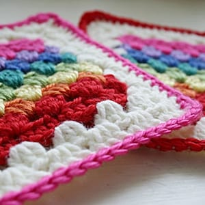 Pode incluir: Dois quadrados de crochê com um padrão de arco-íris em tons de rosa, roxo, azul, verde, amarelo, laranja e vermelho. Os quadrados são enfeitados com crochê branco e rosa.